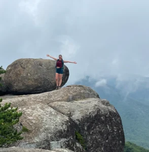 A person with arms outstretched stands on a large rocky ledge next to a massive boulder—perhaps discovering one of the best hikes around Frederick MD. They wear a red shirt and teal shorts, framed by misty mountains and forest in the cloudy background.