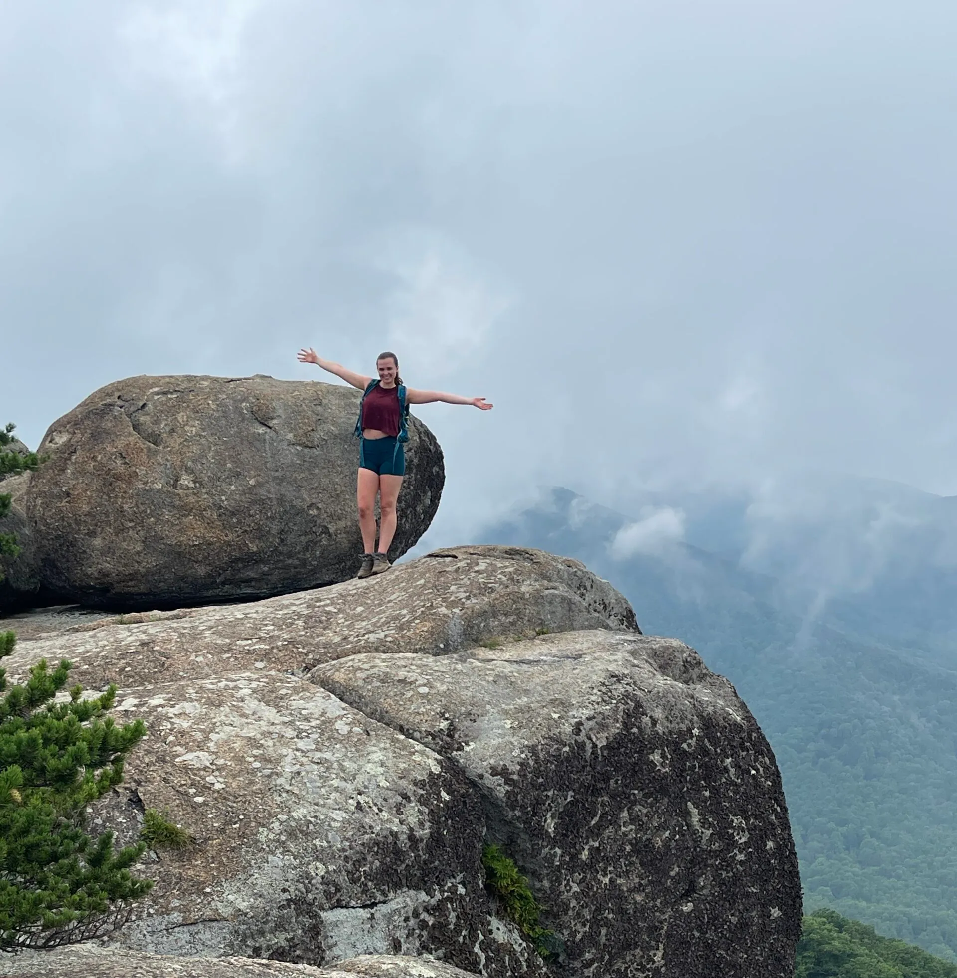 A person with arms outstretched stands on a large rocky ledge next to a massive boulder—perhaps discovering one of the best hikes around Frederick MD. They wear a red shirt and teal shorts, framed by misty mountains and forest in the cloudy background.