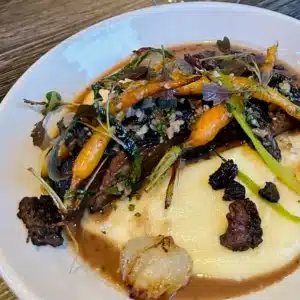 A white plate holds creamy mashed potatoes topped with braised beef, roasted baby carrots, morel mushrooms, and fresh herbs. Inspired by dishes served at the best bars in Frederick, it’s finished with rich brown sauce and microgreens on a wooden table.