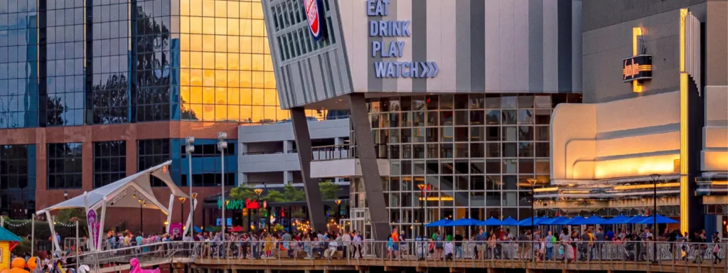 a Busy Waterfront Scene with a Crowd on the Boardwalk Behind Them a Modern Building with Glass Windows and a Large Eat Drink Play Watch Sign Resembles the Vibrant Atmosphere of Frederick Movie Theatre Blue Umbrellas Shade an Outdoor Dining Area