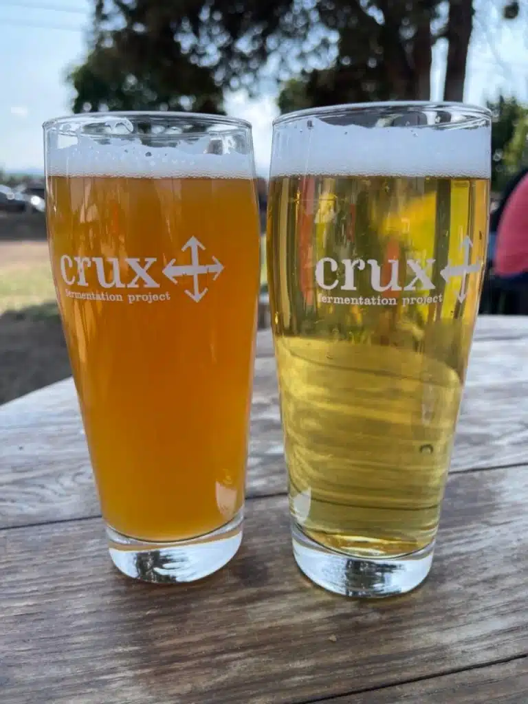 Two pint glasses with Crux Fermentation Project logos sit on a wooden table outdoors in Bend, Oregon. One holds a hazy beer, the other a golden brew—perfect for enjoying one of the best things to do in Bend: relaxing with local craft beer.
