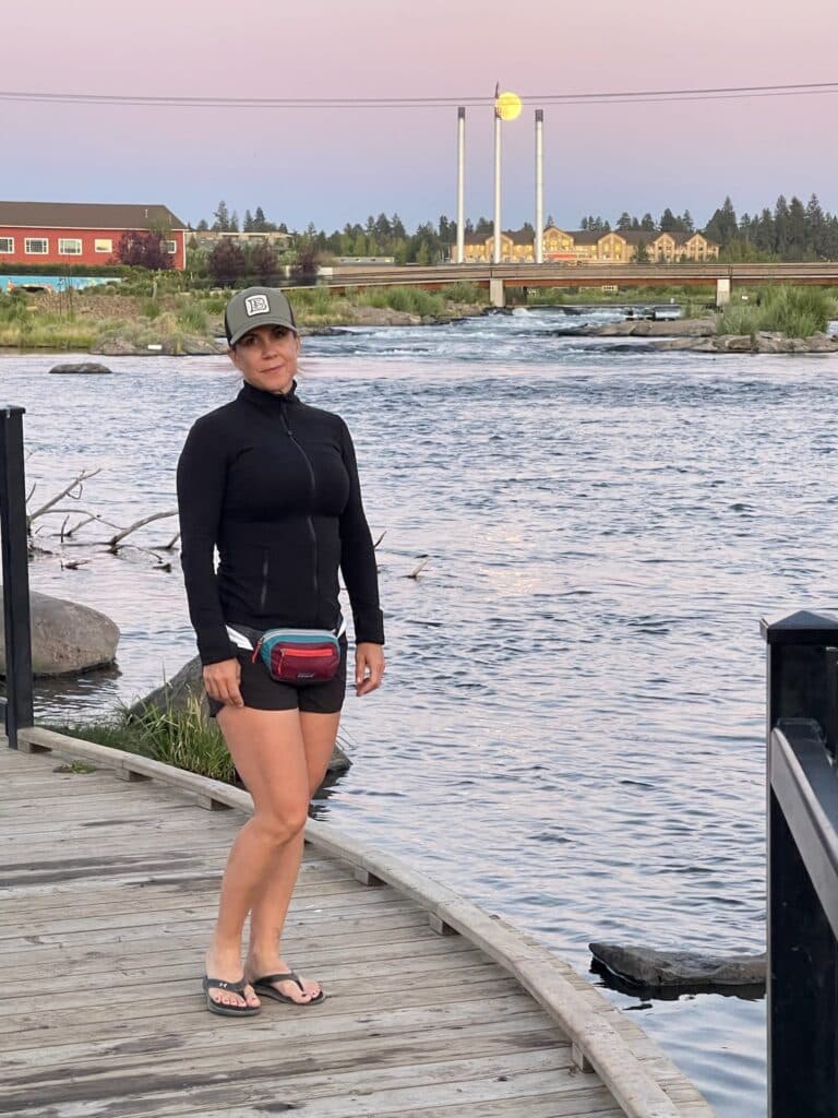 a Woman in a Black Jacket Shorts Cap and Fanny Pack Stands on a Curved Wooden Boardwalk by a Riverone of the Scenic Things to Do in Bend or Behind Her Are Grassy Banks a Red Building and Tall Structures Topped with a Large Yellow Sphere