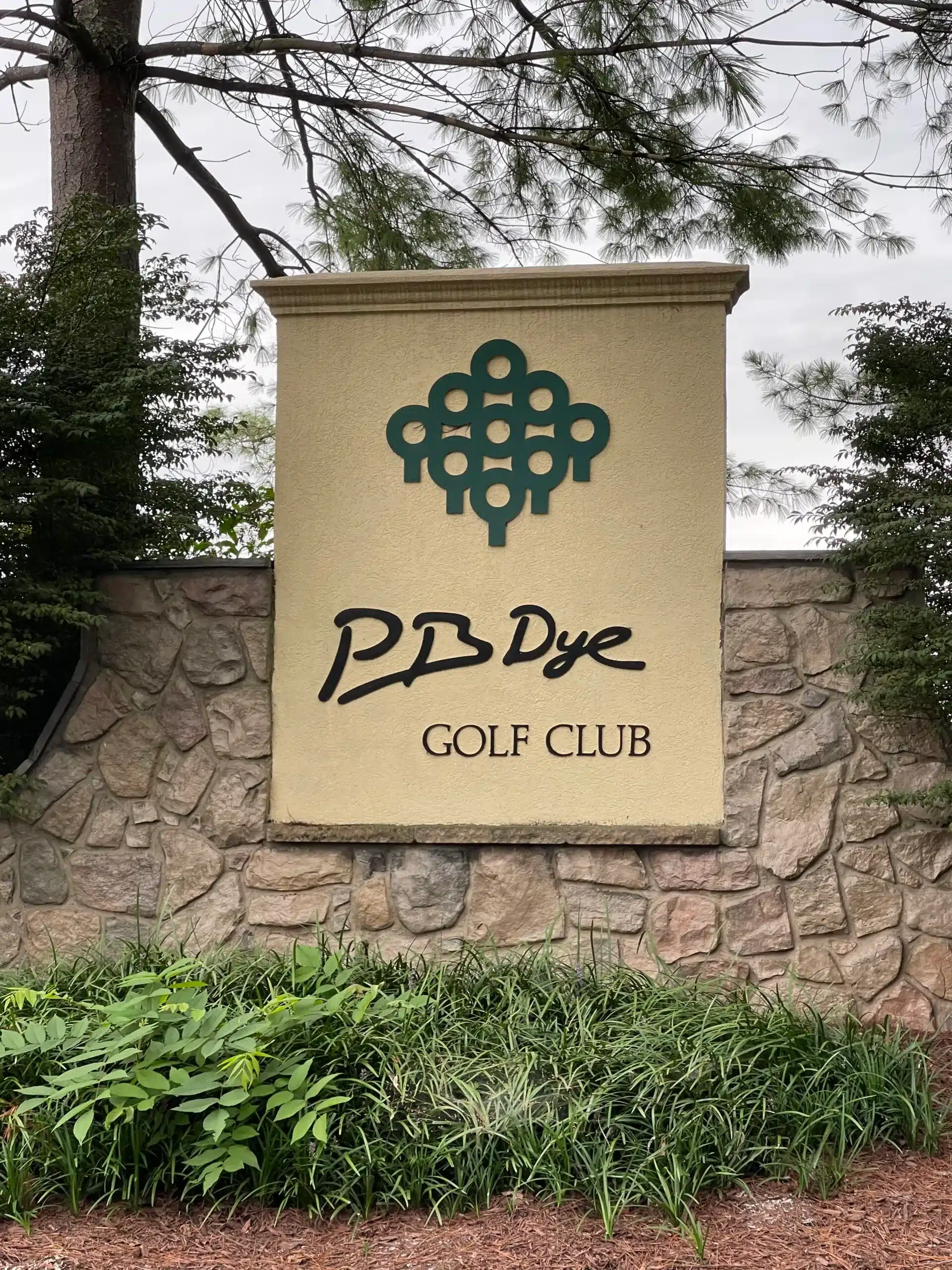 A stone wall displays a beige sign with a green abstract golf ball logo above the words “P.B. Dye Golf Club” in black script. Bushes, tall grass, and trees surround the sign near New Market MD. The sky is overcast.