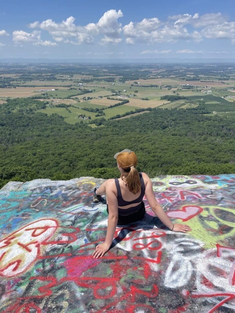 A woman with a ponytail and tan cap sits on a colorful graffiti-covered rock ledge, enjoying one of the best hikes around Frederick MD, as she overlooks green fields and forests under a bright blue sky with scattered clouds.