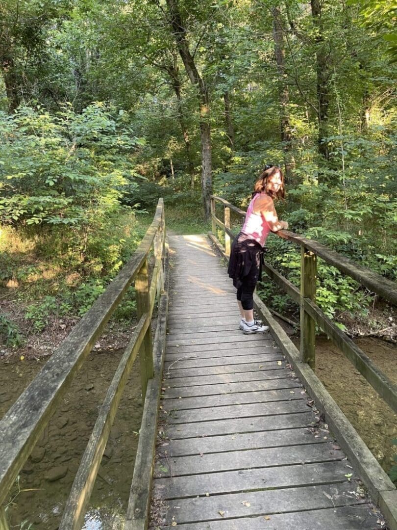 A woman stands on a wooden footbridge in a lush, green forest along one of the best hikes around Frederick MD. She leans on the railing, sunlight filtering through the trees and casting dappled light on the bridge and surrounding foliage.
