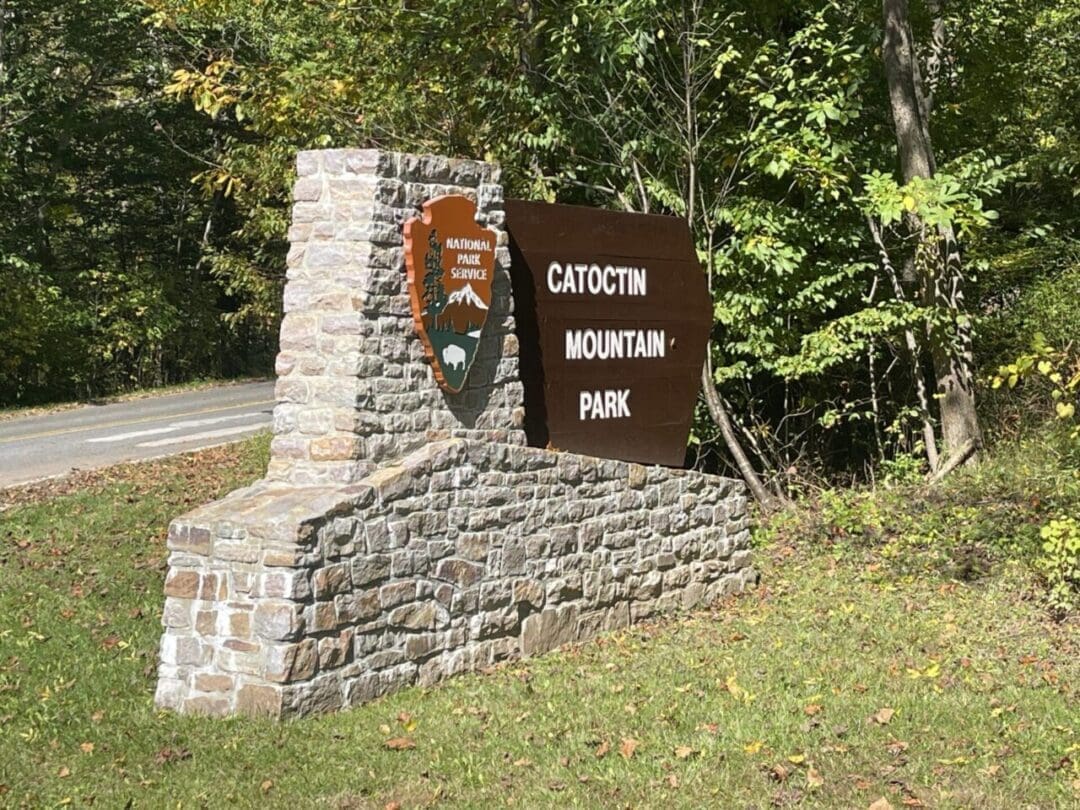 A stone and wood sign at the roadside reads Catoctin Mountain Park with a National Park Service emblem. Green trees and grass surround the sign—an inviting gateway to some of the best hikes around Frederick MD as sunlight filters through the foliage.