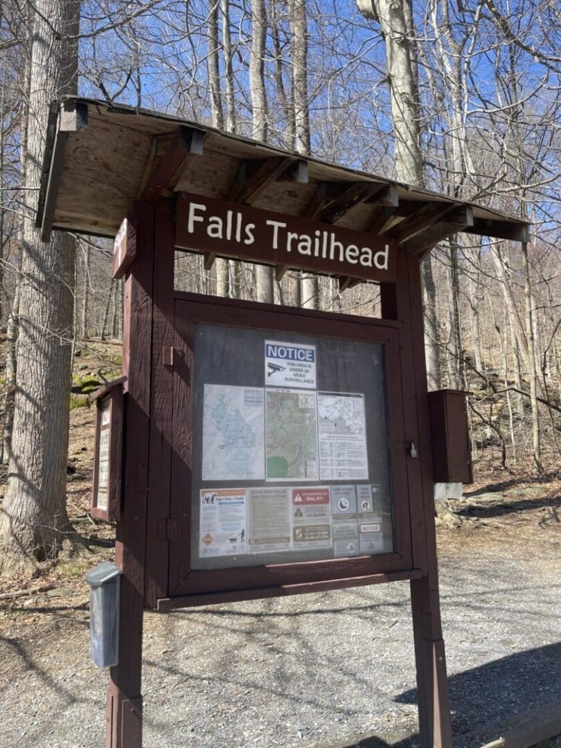 A wooden trailhead sign labeled “Falls Trailhead” stands in a forest with bare trees—one of the best hikes around Frederick MD. The sign displays maps, notices, and safety info inside a glass case as sunlight highlights the gravel-covered ground.