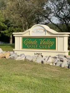 A stone sign with a green and gold plaque reads “Glade Valley Golf Club 10502.” The club’s logo tops it. Surrounded by rocks, grass, and leafy trees under a blue MD sky, it’s one of the scenic golf courses near Frederick.