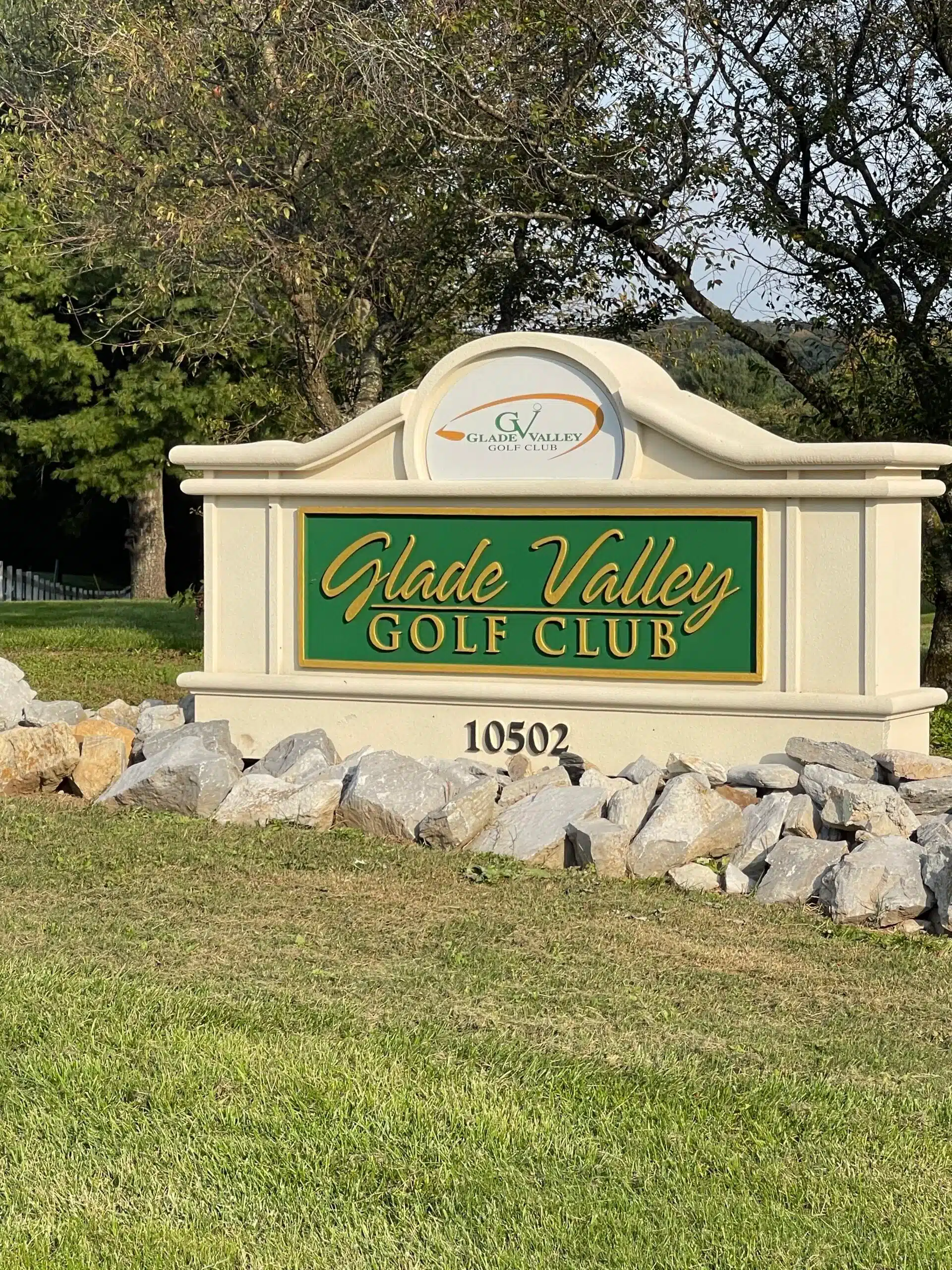 A stone sign with a green and gold plaque reads “Glade Valley Golf Club 10502.” The club’s logo tops it. Surrounded by rocks, grass, and leafy trees under a blue MD sky, it’s one of the scenic golf courses near Frederick.