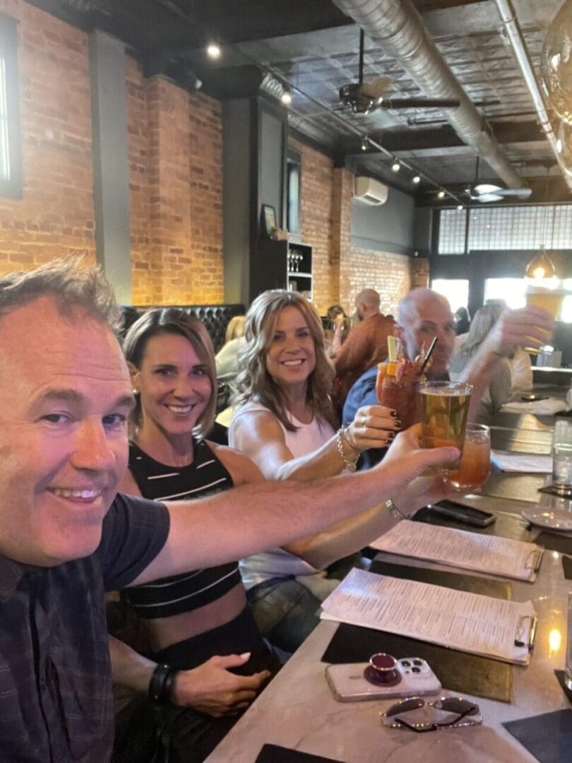 Four Smiling Adults Sit at a Restaurant Table with Exposed Brick Walls Raising Drinks in a Toast Menus and a Phone with a Red Case Rest on the Table Surrounded by the Warm Lighting and Lively Atmosphere of One of the Top Restaurants in Frederick Md