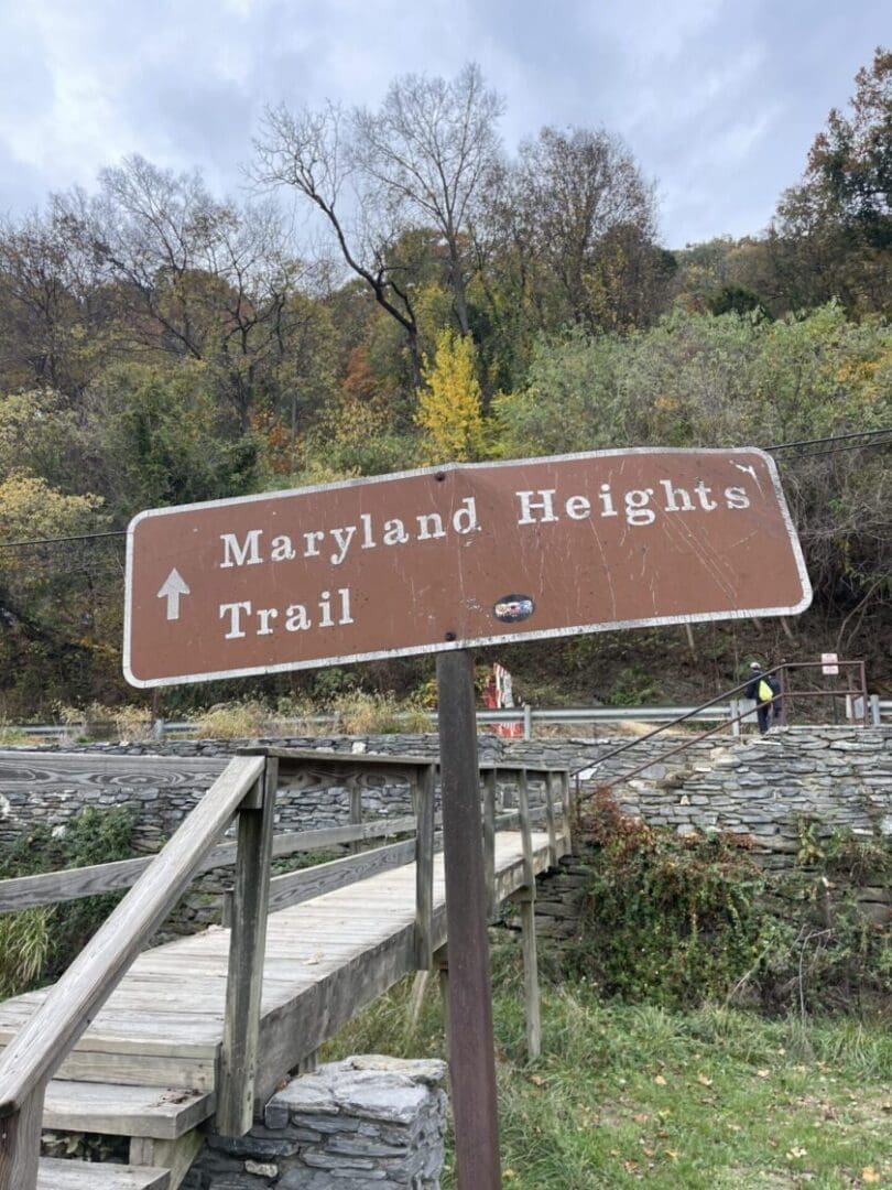 A brown wooden sign reads “Maryland Heights Trail” with a white arrow pointing up. The sign stands beside a wooden footbridge—one of the best hikes around Frederick MD—framed by stone walls and autumn trees under a cloudy sky.