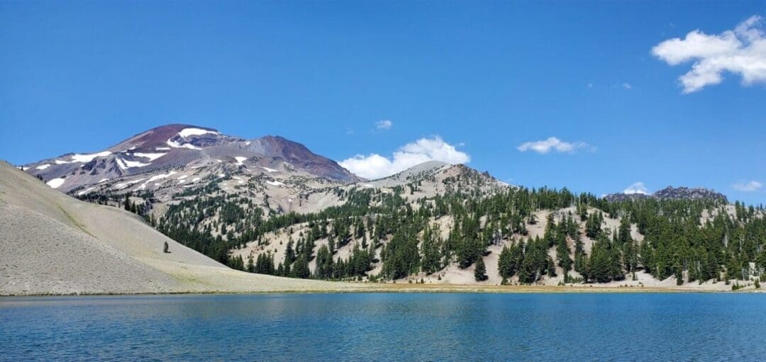 A clear blue lake in the foreground reflects a tree-covered slope; behind it, a rugged mountain with snow patches rises under a bright blue sky—one of the stunning views you can find on Bend hikes.
