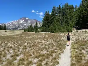 A person walks with arms outstretched on a narrow trail through a grassy field near Bend, Oregon, toward a snow-capped mountain, surrounded by tall green pine trees under a bright blue sky.