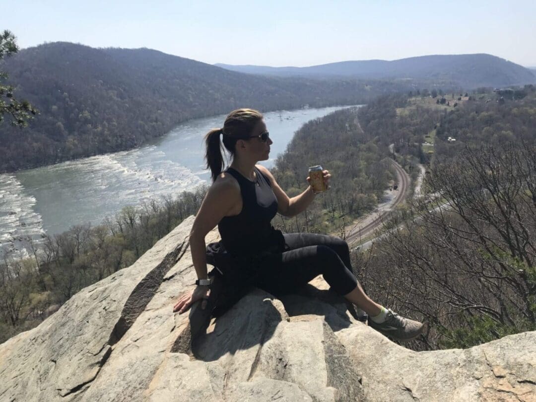 A woman in athletic wear sits on a rocky outcrop overlooking a river and forested hills, enjoying one of the best hikes around Frederick MD. She holds a drink can, wears sunglasses, and gazes at train tracks below under a hazy sky.