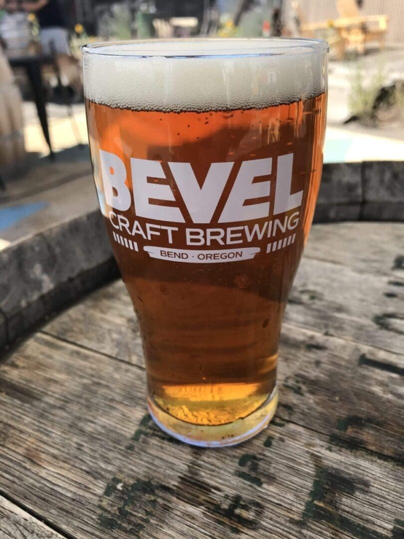 a Pint Glass of Amber Beer from Bevel Craft Brewing One of the Top Bend Breweries Sits on a Weathered Wooden Table Sunlight and Patio Furniture Blur in the Background Capturing a Classic Outdoor Bend Oregon Vibe