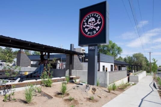 an Outdoor View of a Modern Pub Monkeybar Pub with a Skull Logo the Patio Seating Sits Behind a Gray Wall Surrounded by Sparse Landscaping Under a Sunny Sky Located Among Top Bend Breweries It Faces the Street with a Sidewalk and Utility Poles in Front