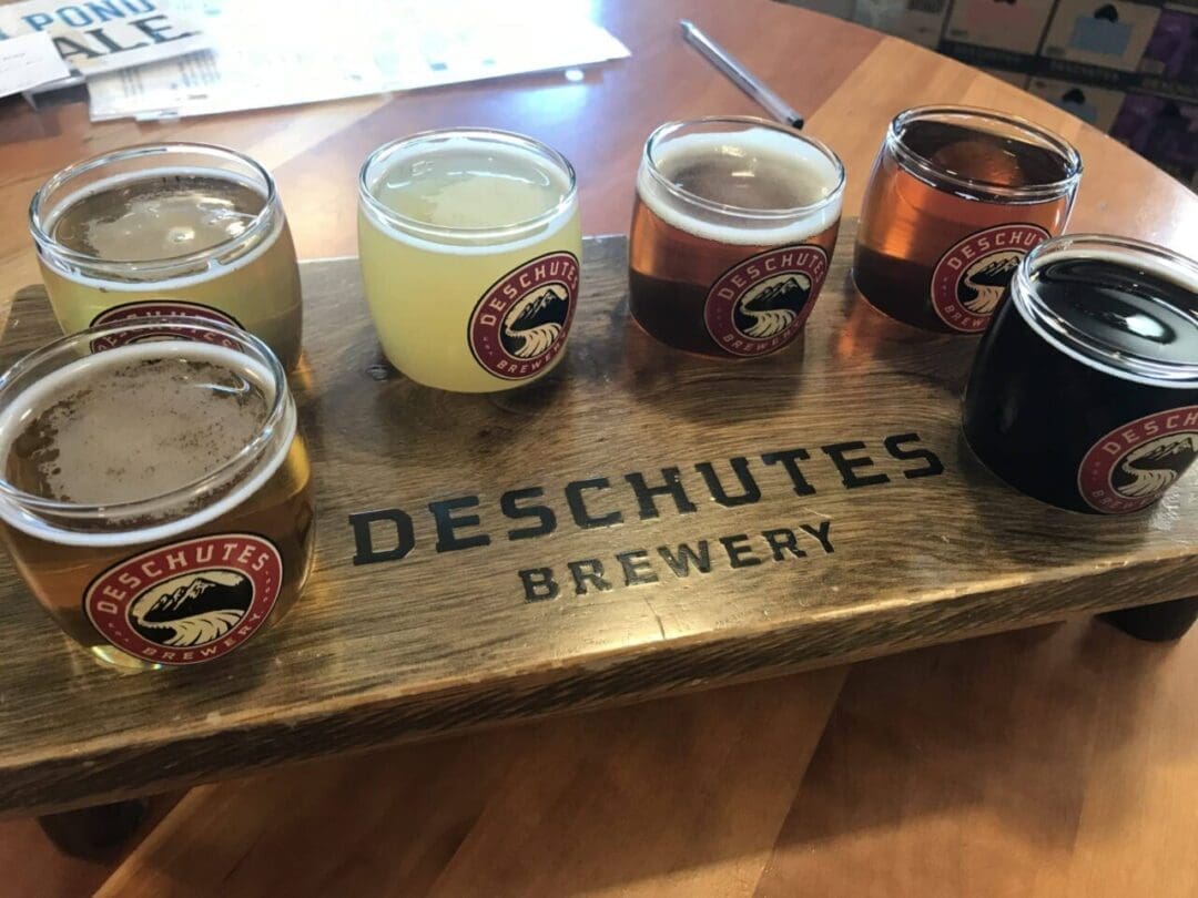 a Wooden Tray Displays Six Small Glasses of Different Colored Beers from One of the Top Breweries in Bend Oregon Each Glass Has the Deschutes Brewery Logo and the Tray Labeled Deschutes Brewery Sits on a Light Brown Wooden Table