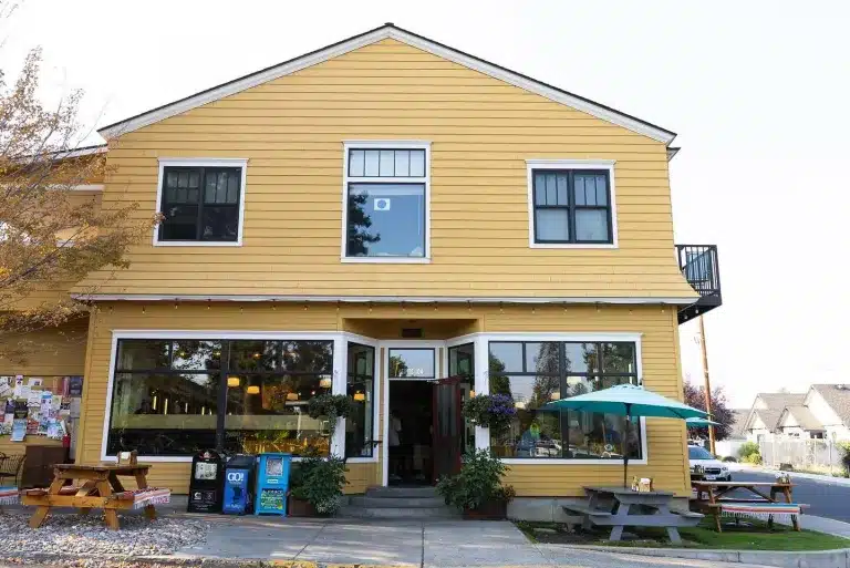 a Two Story Yellow Building with White Trim Houses One of the Best Restaurants Bend Oregon Has to Offer Large Front Windows Hanging Flower Baskets and Outdoor Tables with a Teal Umbrella Invite You in on This Quiet Street