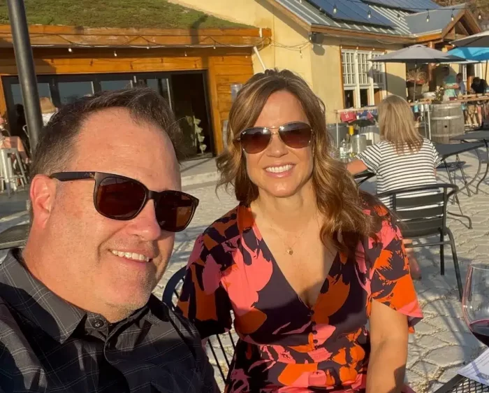 a Man and Woman Sit Outside at a Sunny Patio at One of the Frederick Md Wineries Both Wearing Sunglasses and Smiling the Woman Wears a Colorful Pink and Orange Dress the Man Wears a Dark Plaid Shirt with a Rustic Building in the Background