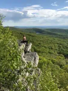 A person sits on the edge of a rocky cliff overlooking a vast green forest, one of the best hikes around Frederick MD. Trees cover rolling hills to the horizon, with a small distant lake nestled in the landscape under a partly cloudy sky.