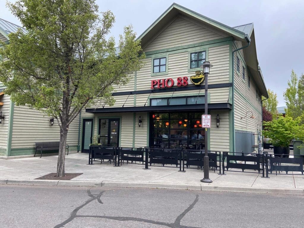 a Light Green Two Story Building Houses Pho 88 a Vietnamese Restaurant Known Among Bend Oregon Restaurants Its Sign Features a Pho Bowl Graphic Outdoor Seating Lines the Sidewalk with a Tree and Streetlamp Nearby Under an Overcast Sky the Street is Empty