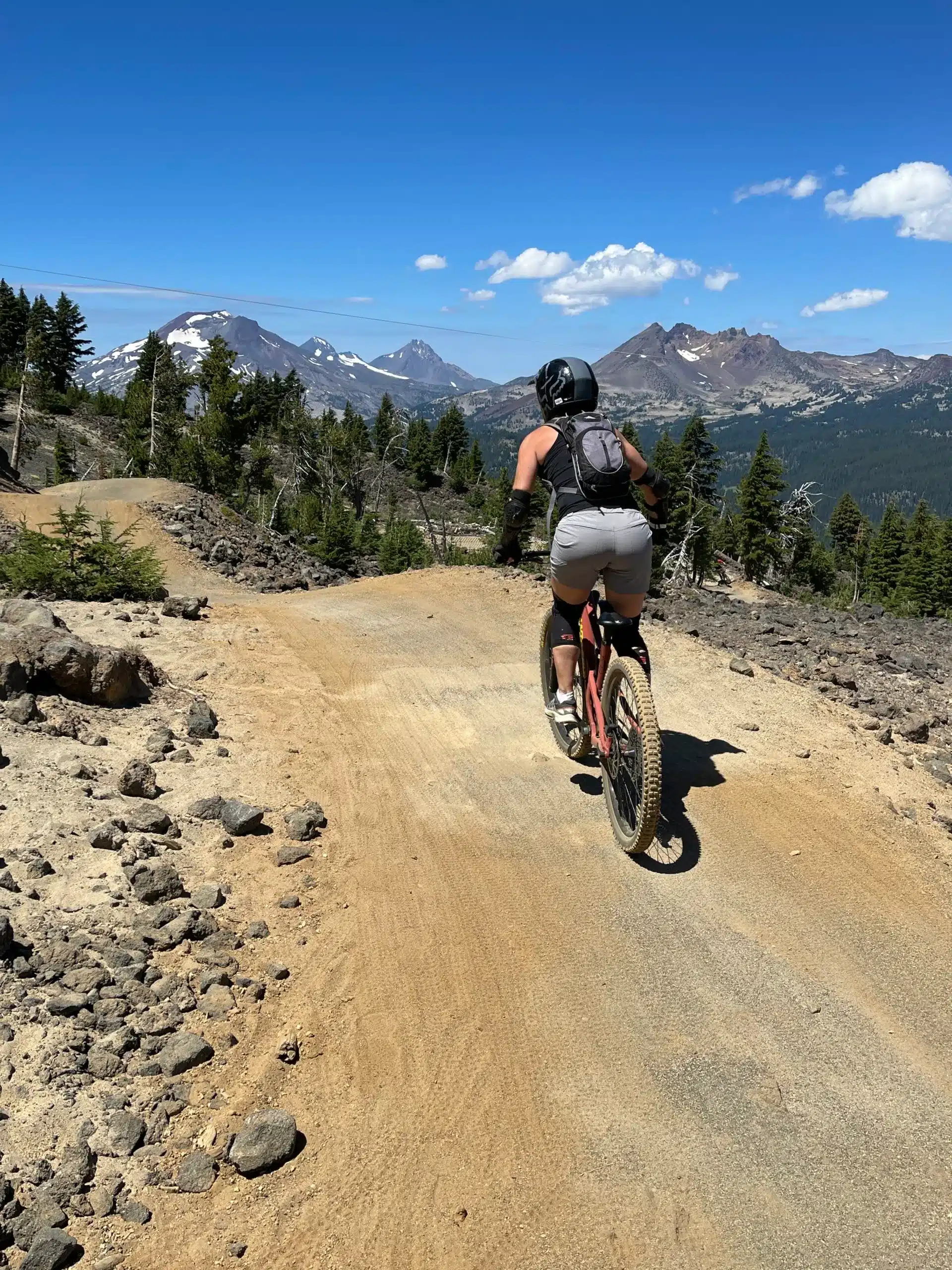 A cyclist wearing a helmet, backpack, and shorts rides a mountain bike up a dirt trail surrounded by rocks and pine trees—one of the top things to do in Bend OR—with snow-capped mountains and a clear blue sky in the background.
