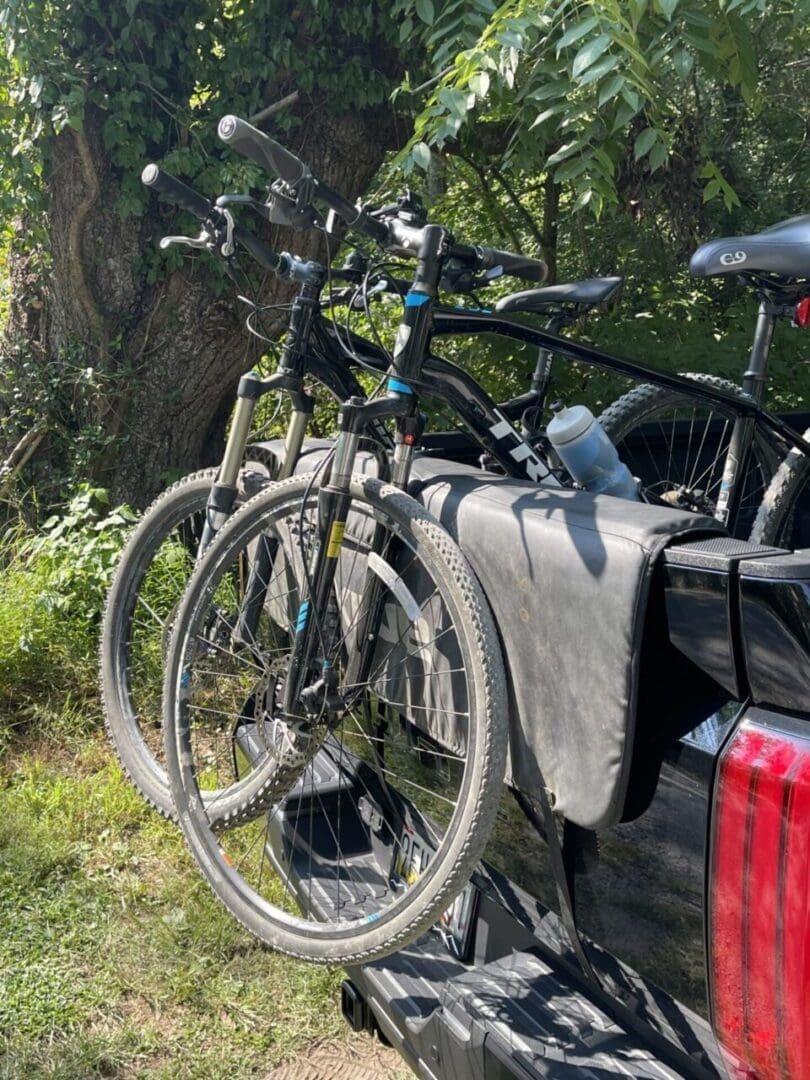 Two black mountain bikes are mounted on the tailgate of a black pickup truck, secured by a padded tailgate cover. Outdoors near Little Bennett Bike Trails, green grass and leafy trees in the background suggest a sunny day.
