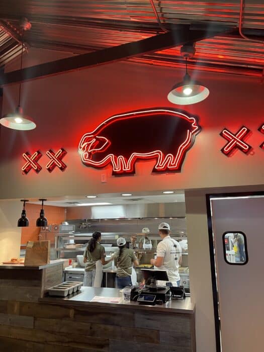 a Neon Red Buffalo Sign and Three Illuminated X Symbols Hang Above an Open Kitchen Where Three People in Casual Clothing and Hats Prepare Food Under Industrial Lighting in a Modern Restaurant Interior Typical of Standout Restaurants in Frederick Md