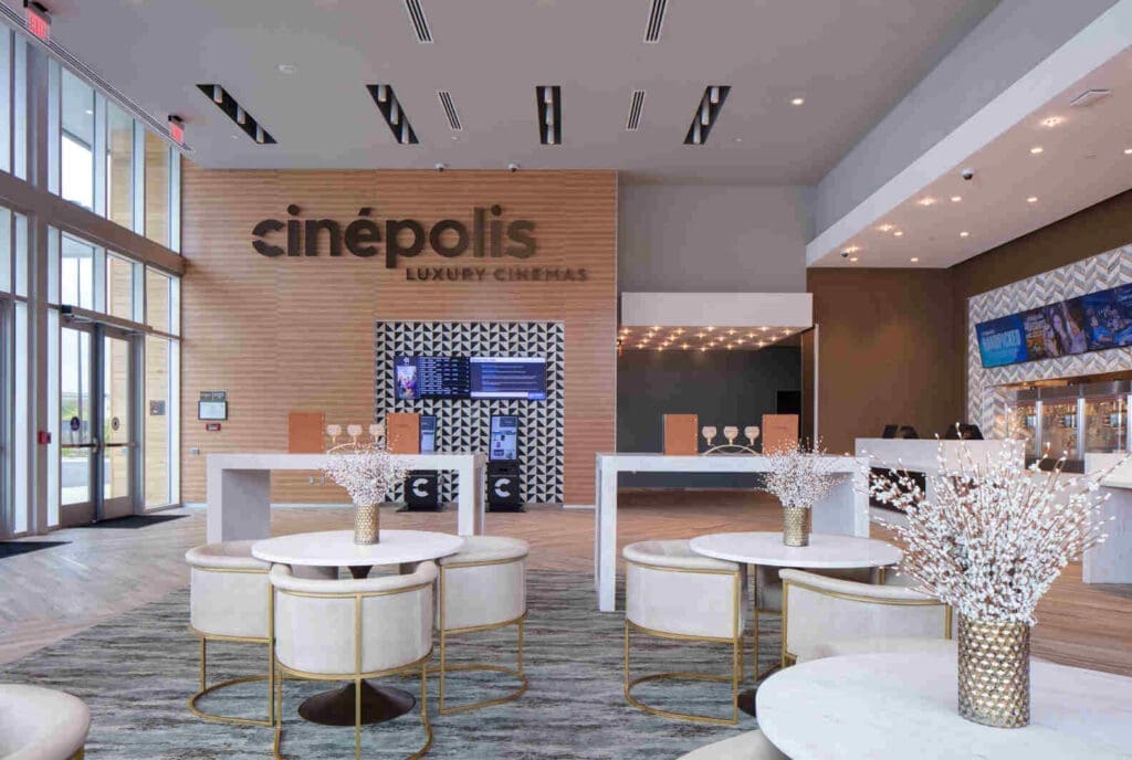 Modern Cinema Lobby at a Frederick Movie Theatre Featuring a Cinépolis Luxury Cinemas Sign on a Wood Accent Wall White Marble Tables and Plush Cream Chairs Fill the Space with Digital Screens Along the Right and Faux Floral Centerpieces on Each Table