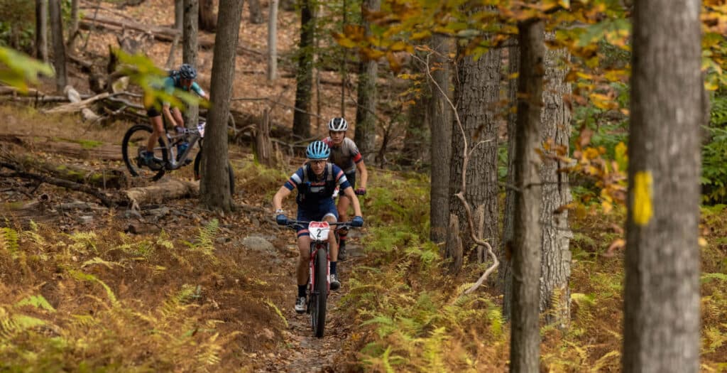 Three Cyclists Ride Mountain Bikes Along a Narrow Dirt Trail Through a Forest with Tall Trees and Autumn Foliage Exploring Bike Trails Near Frederick Md the Lead Rider Wears a Helmet and Number 72 As Sunlight Filters Through the Branches