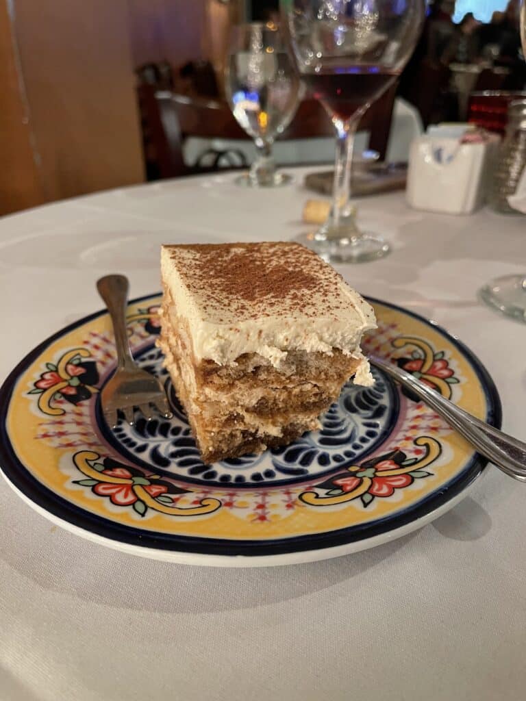 a Slice of Tiramisu Topped with Cocoa Powder Sits on a Colorful Plate a Fork Beside It in the Background Wine Glasses and Table Settings Hint at the Inviting Atmosphere Typical of Frederick Italian Restaurants