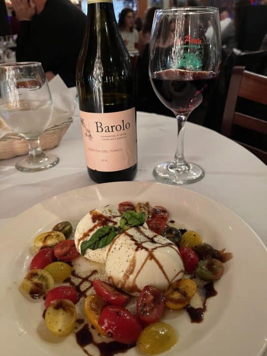 a Plate of Fresh Mozzarella Cherry Tomatoes Basil and Balsamic Glaze Sits on a White Tableclothan Inviting Scene Often Found in the Best Restaurants in Frederick Md Behind It a Glass of Red Wine and a Barolo Bottle Complete the Softly Lit Setting