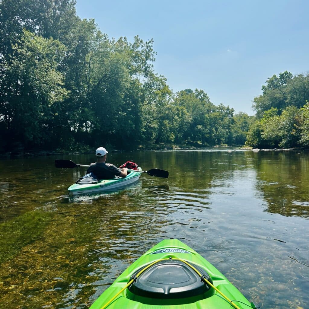 Two People Kayak Down a Calm Shallow River Near Mt Airy Md Surrounded by Lush Green Trees Under a Clear Blue Sky the Photo is Taken from One Kayak Looking Ahead at the Other with Sunlight Reflecting off the Waters Surface
