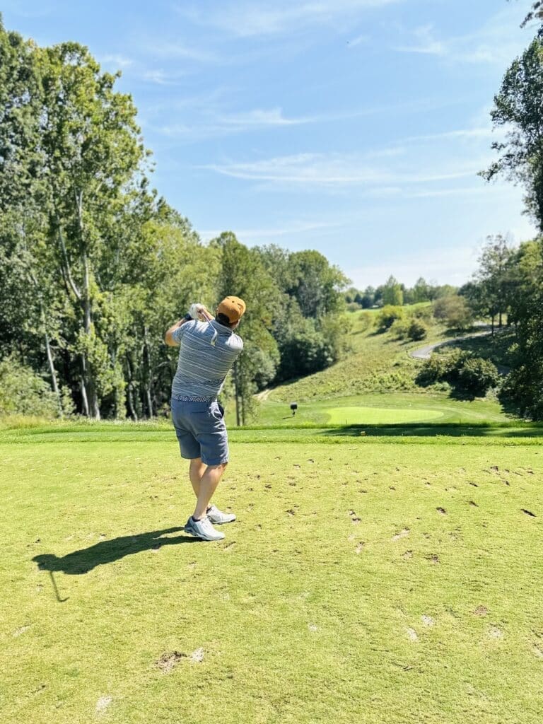 A man in a striped shirt, shorts, sneakers, and an orange cap swings a golf club on a green course surrounded by tall trees and rolling hills. Enjoy scenes like this at golf courses near Frederick, MD under the clear blue sky.