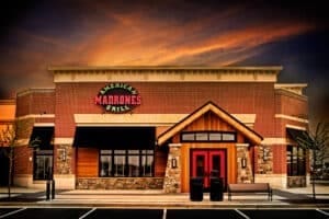 a Red Brick Restaurant with Madrones American Grill Sign Vivid Red Double Doors Stone Details and Peaked Wooden Awning Known As the Best Steakhouse Frederick Md It Sits Beneath a Dramatic Sunset Sky with an Empty Bench and Bike Rack Outside