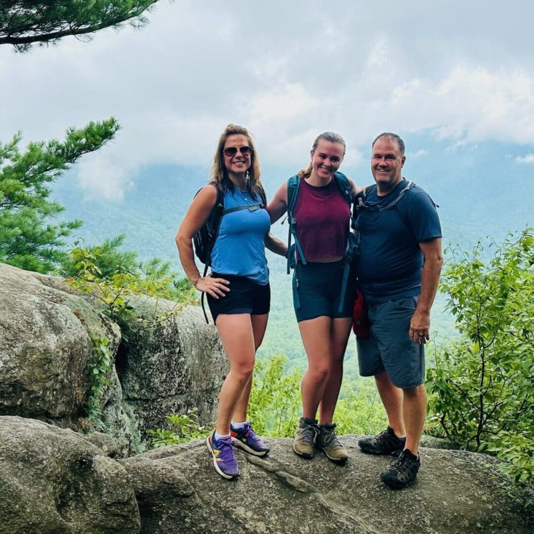 Three people, two women and one man, stand smiling on a large rock surrounded by green trees, ready to explore some of the best hikes around Frederick MD. A misty, mountainous landscape appears in the background under a cloudy sky.
