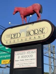 a Restaurant Sign Features a Large Red Horse Statue on Top the Sign Reads the Red Horse Restaurant Promoting Grilled Steaks Seafood Special Events and over 35 Years As the Best Steakhouse Frederick Md Has to Offer Road Signs and Power Lines Are in the Background