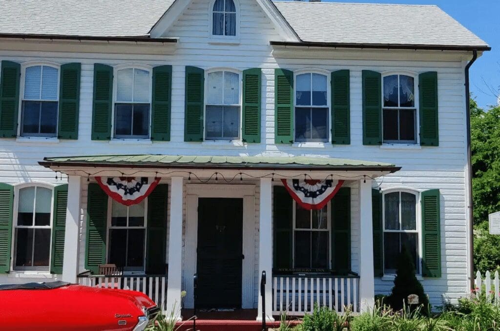A white two-story house with green shutters, a metal porch roof, and patriotic red, white, and blue bunting above the entrance evokes classic charm in New Market, MD. A red car is partially visible by shrubs and a white picket fence lining the porch.