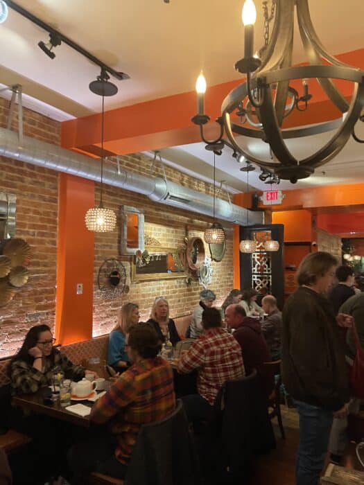 a Warmly Lit Restaurant with Exposed Brick Walls Modern Light Fixtures and Decorative Metal Wall Art Like Many Top Restaurants in Frederick Md Groups of People Dine and Chat While Others Mingle Creating a Lively and Cozy Atmosphere