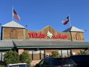a Texas Roadhouse Restaurant Known As the Best Steakhouse Frederick Md with an American Flag on the Left and a Texas Flag on the Right the Wood Sided Building Features Large Windows a Red and White Sign and Cars Parked out Front Under a Clear Blue Sky