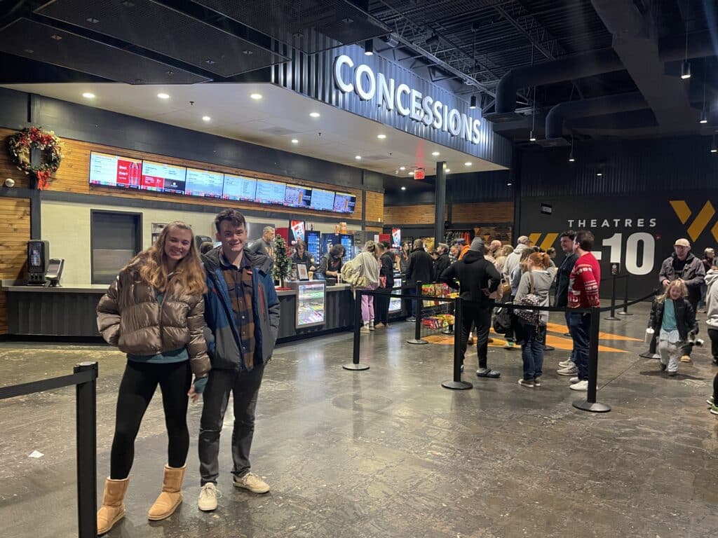 Two Young People Smile and Pose in Front of a Frederick Movie Theatre Concession Stand Behind Them Several People Wait in Line at the Counter Beneath a Large Concessions Sign with Theater Screens and Signs Visible in the Background