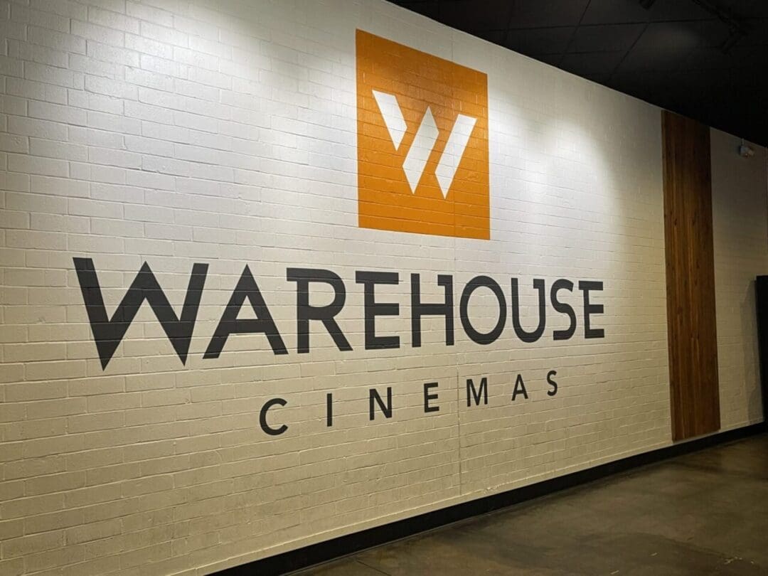 A large white brick wall features the bold black words WAREHOUSE CINEMAS and an orange square above with a white stylized W logo. The polished concrete floor and wood paneling create a modern vibe—one of the top things to do in Frederick, MD.