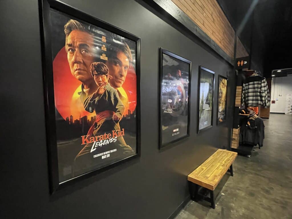 Framed Movie Posters from the Frederick Movie Theatre Hang on a Dark Hallway Wall the Nearest Poster is for Karate Kid Legends Showing Three Serious Faces and Martial Arts Poses a Wooden Bench Sits Below and Shelves on the Right Hold Clothing