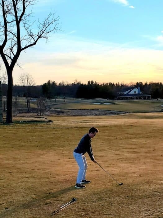 A person stands on a golf course in MD in the late afternoon, preparing to swing a club. The sky is partly cloudy with orange and blue hues. A large tree is left, and a clubhouse with a porch is visible—like golf courses near Frederick.