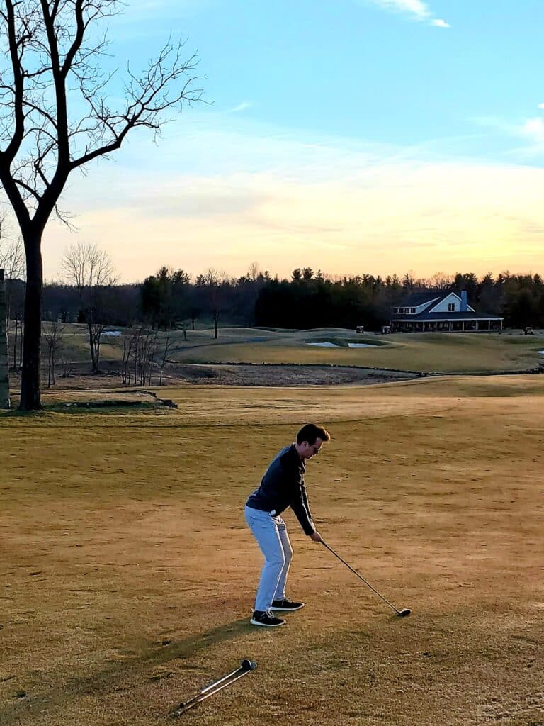 A person stands on a golf course in MD in the late afternoon, preparing to swing a club. The sky is partly cloudy with orange and blue hues. A large tree is left, and a clubhouse with a porch is visible—like golf courses near Frederick.