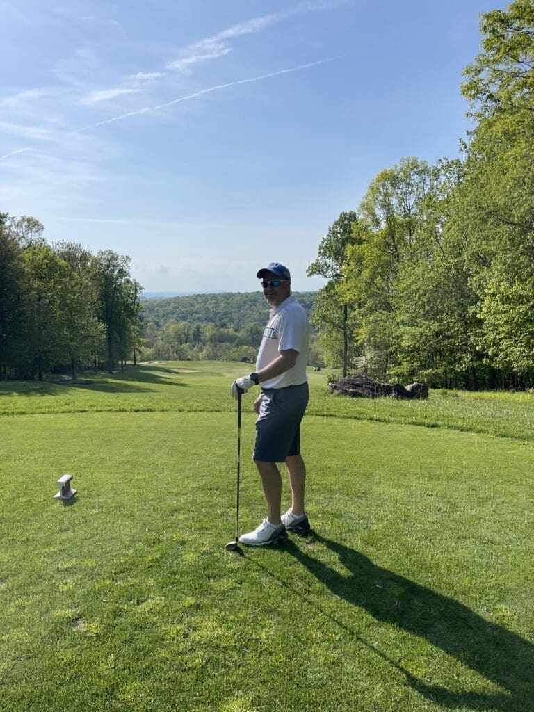 A man in a white shirt, gray shorts, white shoes, and a cap stands on a sunny golf course near Frederick, MD, holding a golf club. He faces the camera and smiles, surrounded by green grass, trees, and distant hills under a clear blue sky.