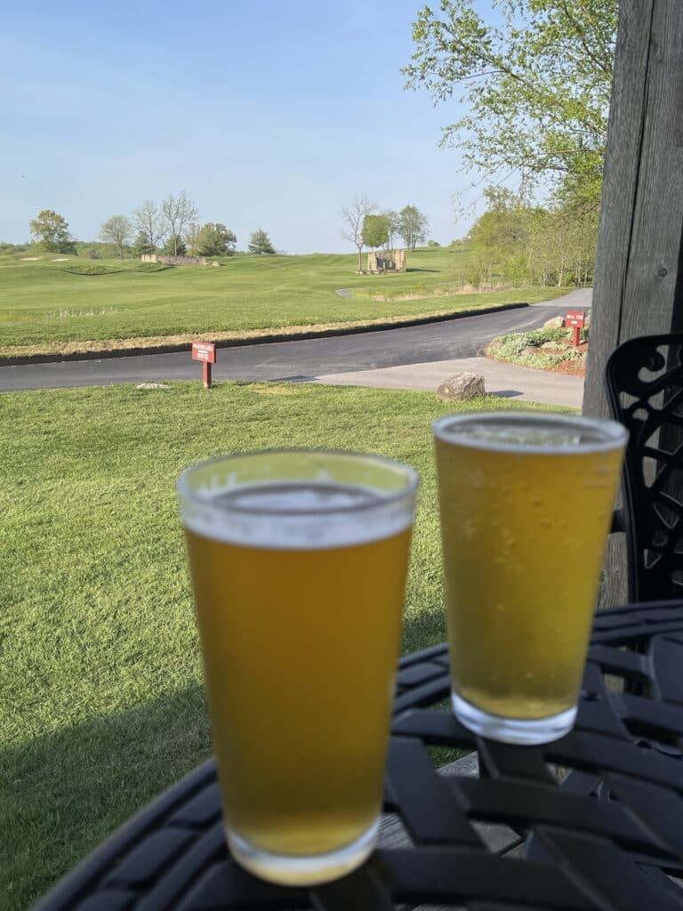 Two glasses of light beer sit on a black metal table outdoors, overlooking green grass and trees at a golf course in MD. The clear blue sky and paved path create a relaxing scene, perfect for enjoying golf courses near Frederick.