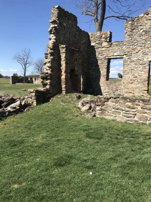 Partially collapsed stone building ruins stand on a grassy field under a blue MD sky. The structure has jagged edges, missing roof, and large open windows. Leafless trees grow behind the building, with another ruin visible near golf courses near Frederick.