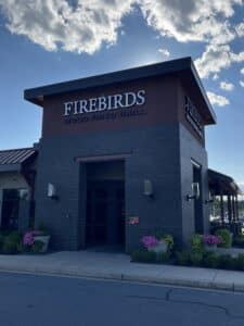 a Modern Dark Brick Restaurant with Large Firebirds Wood Fired Grill Signs on Two Sides This Spot is Known As the Best Steakhouse Frederick Md Offers Small Planters with Pink Flowers Surround the Building Under a Blue Sky a Sidewalk Runs in Front