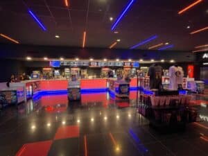 a Modern Frederick Movie Theatre Concession Stand with Bright Blue and Red Neon Lights Counters Display Snacks and Popcorn Machines Merch Tables with T shirts and Baskets Are in Front Set on Glossy Black and Red Tiles the Area is Empty of People